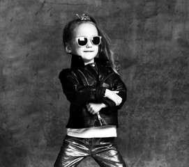 Naklejka premium Black and white portrait of stylish kid girl in leather jacket, shimmering pants and sunglasses is cool posing