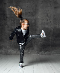 Fototapeta na wymiar Active frolic kid girl in leather jacket, pants, shimmering sneakers and sunglasses is jumping lifting her leg high