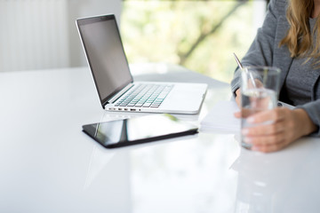 A blonde hair businesswoman working in her workstation. Businesswoman working at the desk at workplace. Woman working in home office. Woman sitting at desk and working with laptop and glass of water.