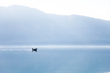 Fototapeta premium Blue background. Boat with fisherman on sea. Fishing in foggy morning lake. Amazing nature landscape with mountains, silence, calmness. Reflection in still water of Kotor Bay, Montenegro. Copy space