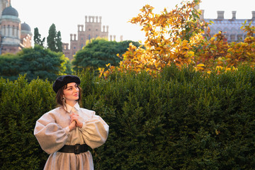 Mysterious young woman in black beret and grey dress holds hands together and looking away, green bushes on the background