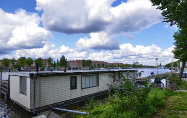 Fototapeta na wymiar Amsterdam, Holland, August 2019. View of the Amstel River, outskirts of the city. A large houseboat moored on the river bank. Sunny day with blue sky and white clouds.