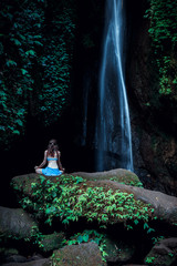 Young woman sitting on the rock, practicing yoga near waterfall. Hands in gyan mudra. Leke Leke waterfall, Bali. View from back.