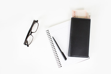 The top view of the pen and the money in a black leather bag placed on a book with eye glasses on a white table. With copy space.