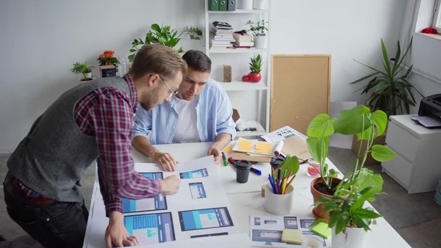 High angle view of two designers examining and discussing website interface samples for different devices printed on paper sheet at desk in office