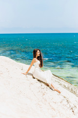 Fototapeta na wymiar Charming girl in a white dress sits on a rock on the background of the sea