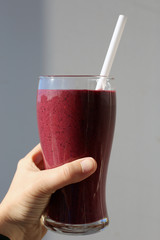Pink / purple homemade berry and vegetable smoothie with vibrant color and plenty of vitamins. Healthy and delicious drink for sunny summer days. Hand holding the smoothie, grey background, sunny day.