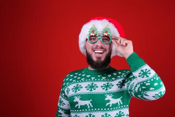 Studio portrait of handsome bearded man wearing christmas sweater with snowflake ornament, posing over the red wall, copy space for text. Festive background. Male with facial hair smiling.