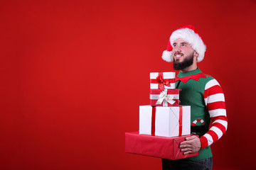 Studio portrait of handsome bearded man wearing traditional elf costume, green vest & striped sleeve, posing over the red wall, copy space for text. Festive background. Male with facial hair smiling.