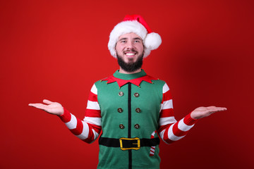 Fototapeta na wymiar Studio portrait of handsome bearded man wearing traditional elf costume, green vest & striped sleeve, posing over the red wall, copy space for text. Festive background. Male with facial hair smiling.