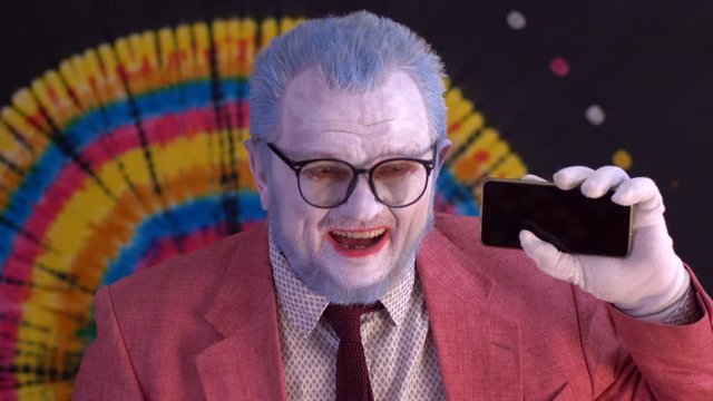 elderly mime man in big glasses with blue hair and beard in bright pink jacket and tie looks at smartphone screen and shows it to audience. Laughter, tears, positive and negative emotions. Happy clown