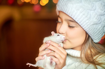 Cute little girl in a hat holds in her arms and hugs a funny white rat on a background of bright holiday lights. Symbol of 2020. Pets