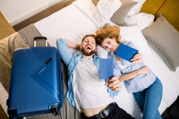 A happy young couple on their vacations lying on the bed in hotel room and holding blue passports. Focus on passports in hands