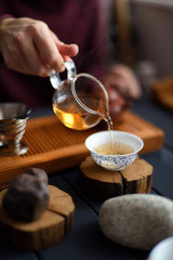 Tea ceremony. Slender woman hand pouring freshly brewed green tea from glass pitcher into Chinese cup and decorative stones on wood slabs on dark background