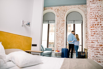 Young cheerful couple embracing, while arrive in a hotel room with big suitcases, back view