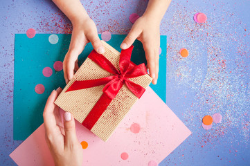 Woman's and child's hands holding a giftbox tied with bright red color ribbon on purple background with sparklets and confetti.