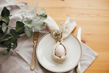 Stylish Easter brunch table setting with egg in easter bunny napkin on  table. Modern natural dyed pink easter egg on napkin with bunny ears, flowers on plate and cutlery. Easter table decorations.