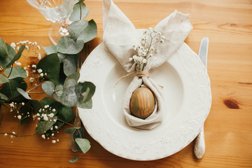 Stylish Easter brunch table setting with egg in easter bunny napkin. Modern natural dyed green egg on napkin with bunny ears, flowers on vintage plate and cutlery. Easter table decorations