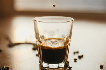 Coffee drops close up in warm light while pouring hot espresso in glass cup on background of roasted coffee beans on wooden table. Alternative Coffee brewing concept. Copy space