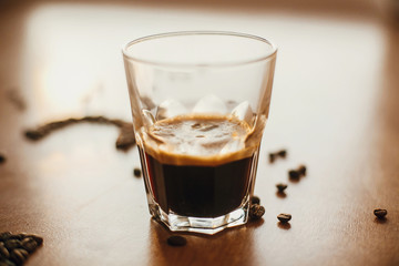 Hot espresso with foam in glass cup with aromatic roasted coffee beans on wooden table in warm light.  Alternative Coffee brewing concept. Copy space