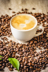 Cup of coffee with foam on the scattered grains on the burlap. Fragrant invigorating drink from freshly roasted beans.