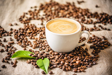 Cup of coffee with foam on the scattered grains on the burlap. Fragrant invigorating drink from freshly roasted beans.