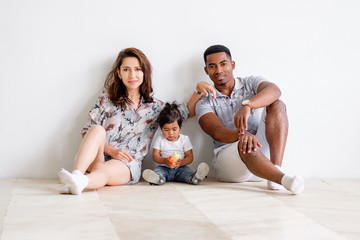 Loving young woman is looking at her young african-american husband while sitting on floor with her little charming mixed-race daughter. Concept of interracial american happy family. Advertising space
