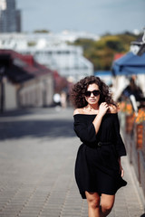 Fototapeta premium Beautiful curly brunette woman in sunglasses and a black dress walks around the city on a bright sunny day.