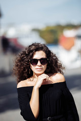 Beautiful curly brunette woman in sunglasses and a black dress walks around the city on a bright sunny day.