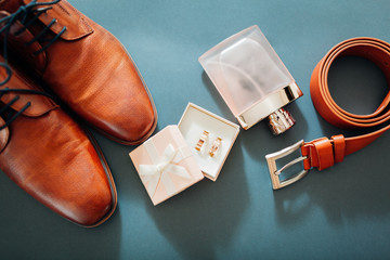 Groom's wedding day accessories. Brown leather shoes, belt, perfume, golden rings on grey background. Male fashion