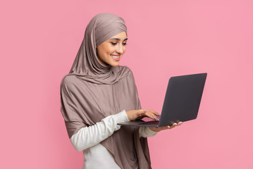 Smiling muslim girl in hijab holding and typing on laptop