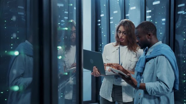 Female IT professional instructing young server technician inside working server cabinet. Male and female server engineers coworking with laptop inspecting data center cyber security.