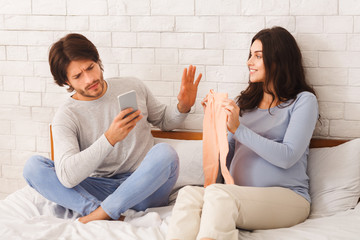 Indifferent husband ignoring his pregnant wife, using smartphone in bedroom