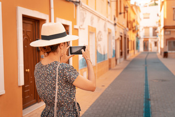 Happy tourist woman with straw sunhat taking a picture of a narrow and picturesque street in Denia, Alicante province, Spain. , Alicante, Spain. Teal and orange style.