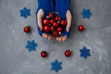 Hands of young beautiful woman in blue mittens. She holds Christmas red balls. Nearby are blue snowflakes and red decorations on the Christmas tree.