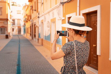 Happy tourist woman with straw sunhat taking a picture of a narrow and picturesque street in Denia, Alicante province, Spain. , Alicante, Spain. Teal and orange style.