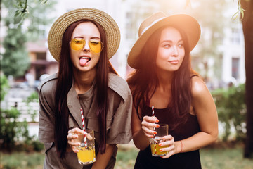 Two cute brunette girlfriends in straw hats are sitting in the park, have a summer picnic in the park. girls drink orange juice and eat croissant outdoors. Close Up portrait of girlfriends at sunset.