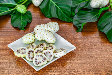 Noni fruits and noni slices on white plate, green leaves and wooden background