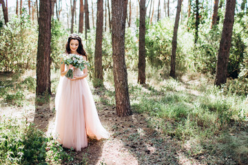 Obraz na płótnie Canvas Beautiful bride in a tender pink dress outdoors. Porter in full growth of a beautiful brunette woman with long dark hair. Girl holds a bouquet. A girl in a long dress walks in a pine forest.