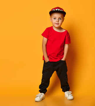 Young guy boy in a red T-shirt and dark pants, white sneakers and a funny cap posing on a free copy space on a yellow background
