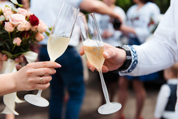 The bride and groom are toasting with their festive champagne glasses outside, close up. Blurred bouquet, and guests on background