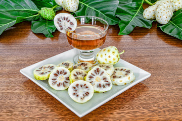Noni fruits and noni slices on white plate, juice in a glass on green leaves and wooden background