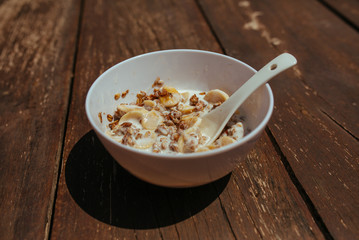 Muesli with banana in white bowl with spoon on a wooden bench