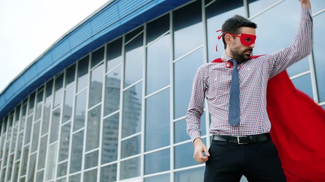 Portrait of funny young man wearing cape and mask making superhero gestures then running to big business rushing to success. People and achievements concept.