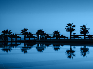 Obraz premium Sunset with silhuettes palm and reflection in swimming pool. Blue color background.