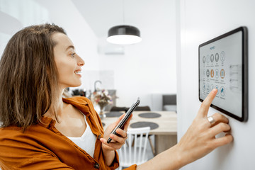 Young woman controlling home with a digital touch screen panel installed on the wall in the living...