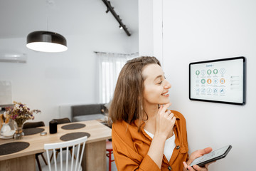 Young woman controlling home with a digital touch screen panel installed on the wall in the living...