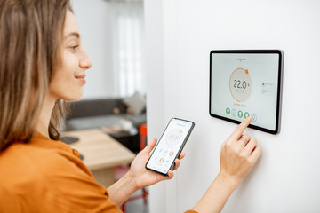 Young woman controlling temperature in the living room with smart phone and digital touch screen...