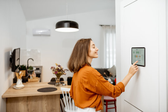 Young woman controlling home with a digital touch screen panel installed on the wall in the living room. Concept of a smart home and mobile application for managing smart devices at home