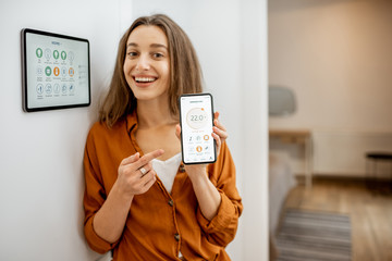 Portrait of a young and happy woman near a touch screen panel for managing smart home. Holding...